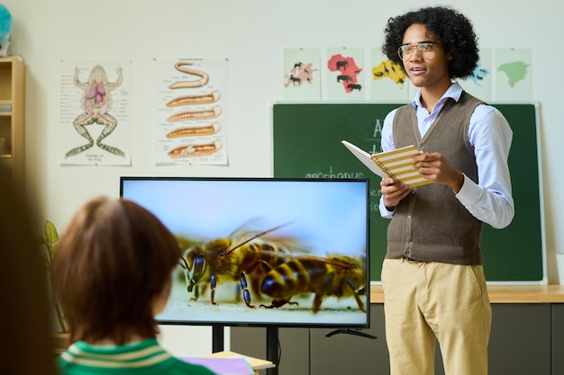 Chico afroamericano con cuaderno de pie junto a una pantalla interactiva con abejas