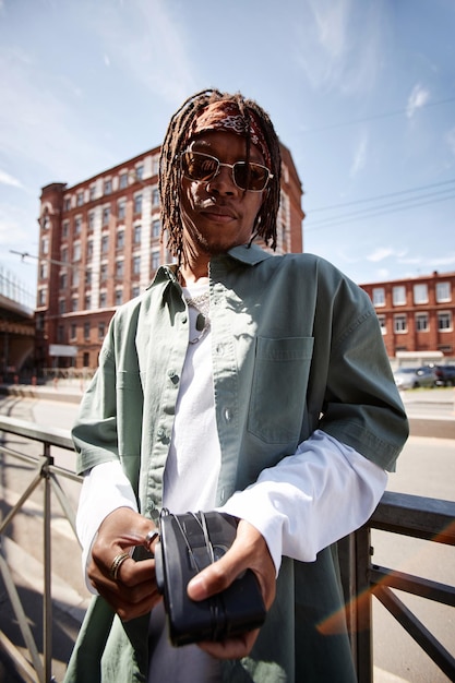Chico afroamericano elegante con rastas y cinta para la cabeza mirando a la cámara mientras está de pie en un entorno urbano contra las barandillas