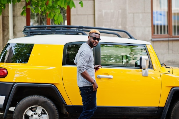 Chico afroamericano con estilo en suéter gris y gafas de sol negras posadas en la calle contra el coche amarillo