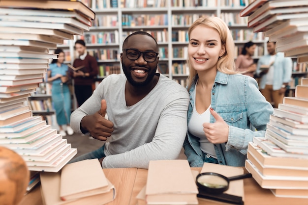 Foto chico afroamericano étnico y chica blanca rodeada de libros en la biblioteca. los estudiantes están dando el visto bueno.