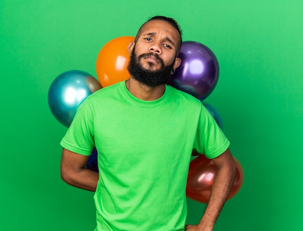 Chico afroamericano joven disgustado con camiseta verde de pie delante de globos poniendo la mano en la cadera aislada en la pared verde