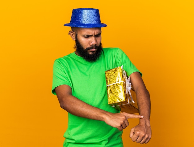 Chico afroamericano joven disgustado con gorro de fiesta sosteniendo una caja de regalo que muestra el reloj de pulsera