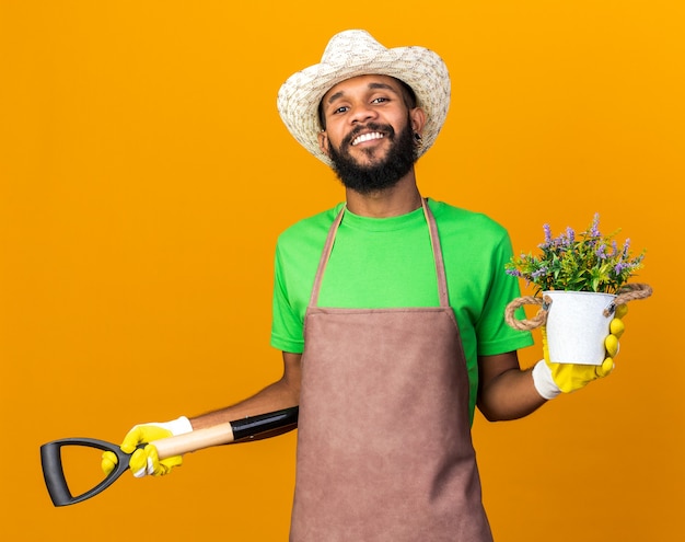 Chico afroamericano joven jardinero confiado con sombrero de jardinería y guantes sosteniendo la pala con flor en maceta