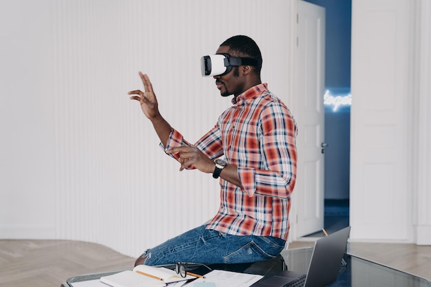 Un chico afroamericano moderno con gafas VR interactúa con la realidad virtual sentado en el escritorio de la oficina