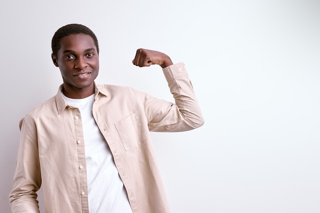 Chico afroamericano mostrando los músculos del brazo, hombre fuerte y poderoso aislado en la pared blanca del estudio
