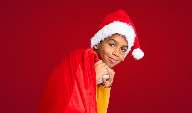 Chico afroamericano con sombrero de navidad y llevar una bolsa con regalos sobre fondo rojo.