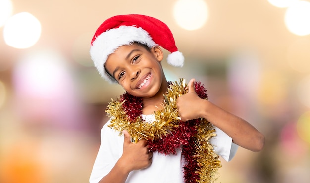 Chico afroamericano con sombrero de Navidad con pulgares arriba porque algo bueno ha sucedido sobre fondo borroso