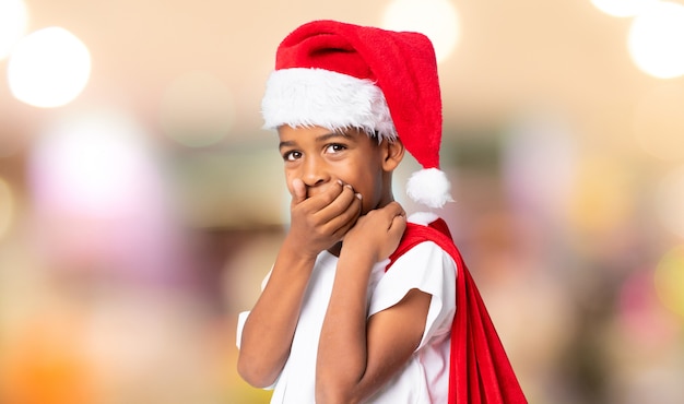 Chico afroamericano con sombrero de navidad y tomar una bolsa con regalos haciendo gesto de sorpresa sobre fondo borroso