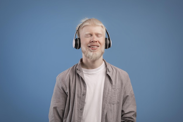 Chico albino tranquilo satisfecho en auriculares disfrutando de su música favorita con los ojos cerrados de pie sobre azul