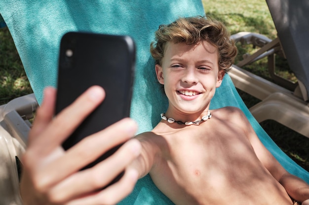 Chico alegre de cabello rubio sonriendo y mirando la pantalla del teléfono móvil mientras toma selfie en una silla de manta en el jardín del complejo
