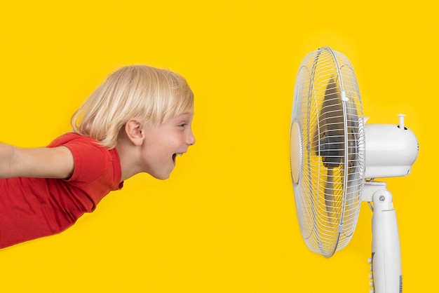 Chico alegre con cabello rubio vuela al ventilador Concepto de calor de verano Fondo amarillo brillante
