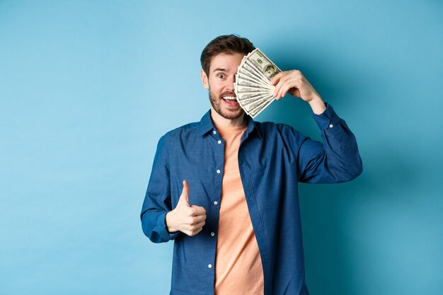 Chico alegre cubre la mitad de la cara con dinero y mostrando el pulgar hacia arriba, recomendando un préstamo en efectivo rápido, de pie sobre fondo azul.