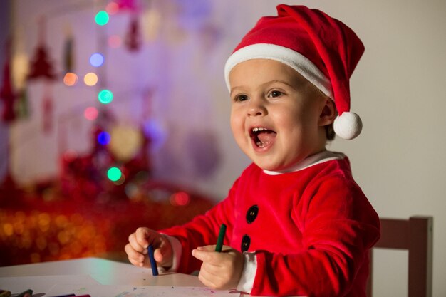 Un chico alegre disfrazado de Papá Noel sentado a la mesa con lápices mientras sonríe y mira hacia otro lado en un apartamento con un fondo borroso