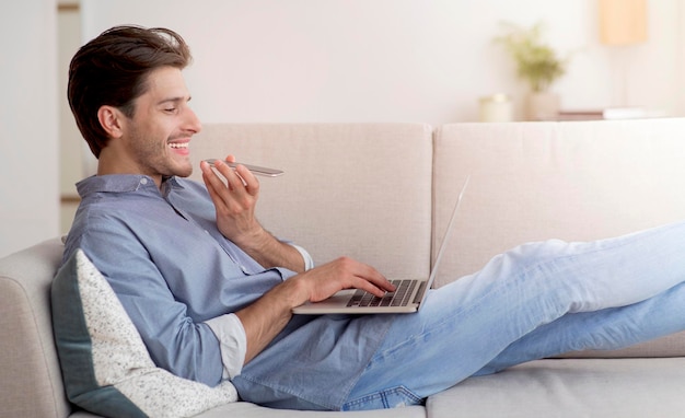 Chico alegre hablando por teléfono usando una computadora portátil acostado en el sofá