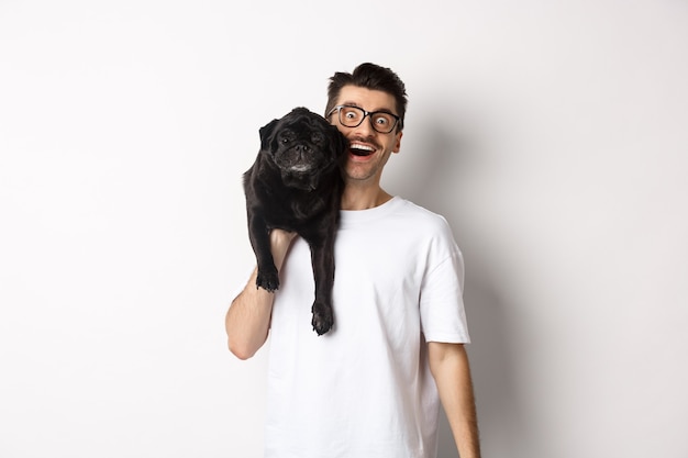 Chico alegre hipster con gafas y camiseta, lleva lindo pug negro en el hombro y sonriendo. Dueño de perro jugando con su mascota, de pie sobre fondo blanco.