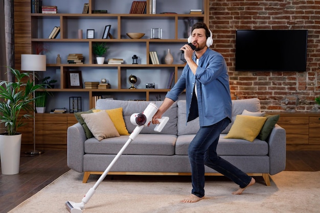 Chico alegre, el hombre limpia el apartamento Pasa la aspiradora usando auriculares bailando y cantando en un teléfono inteligente en lugar de un micrófono La limpieza de la casa es fácil