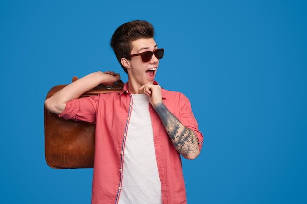 Chico alegre joven inconformista con brazo tatuado en gafas de sol negras y camisa roja con bolsa y mirando a otro lado con la boca abierta contra el fondo azul