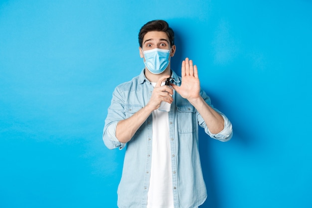 Chico alegre en máscara médica que muestra cómo desinfectar las manos con desinfectante, usando antiséptico, previniendo la propagación del virus, pared azul