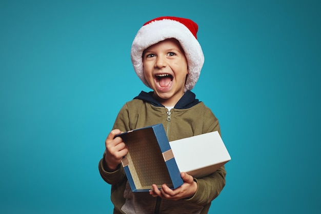 Chico alegre niño con sombrero de navidad abriendo una caja de regalo aislada sobre azul