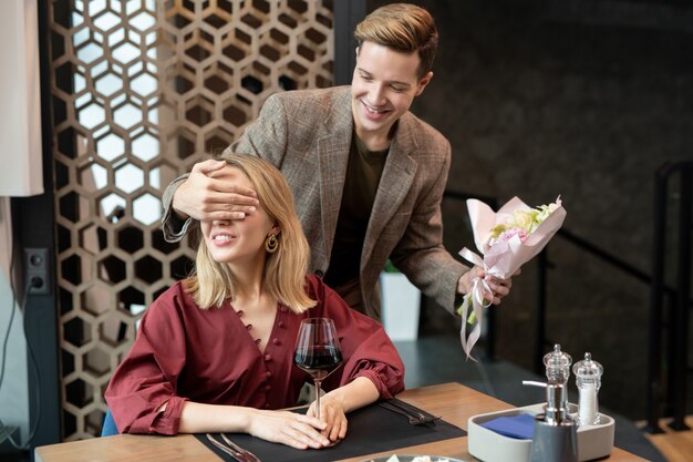 Chico alegre con ramo de flores cubriendo los ojos de su novia con la mano mientras está de pie detrás y haciendo su agradable sorpresa en el restaurante