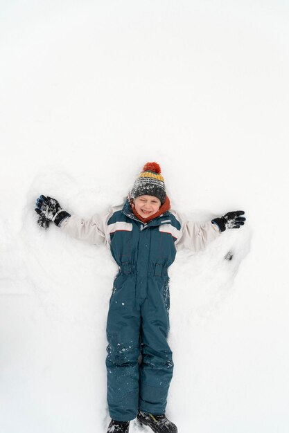 Un chico alegre yace en la nieve y hace un ángel de nieve. Infancia feliz. Vista superior. Marco vertical