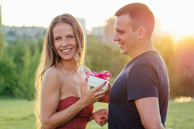 Chico amante da una caja de regalo a avergonzada atractiva hermosa chica rubia de pelo largo enamorada en vestido de cóctel de noche en el parque de verano con fondo de hierba verde. Concepto de San Valentín