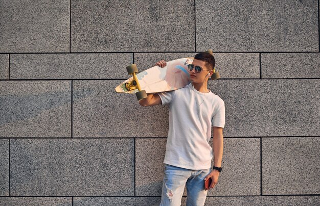 Chico americano caucásico joven con gafas sosteniendo un longboard en su hombro contra la pared gris