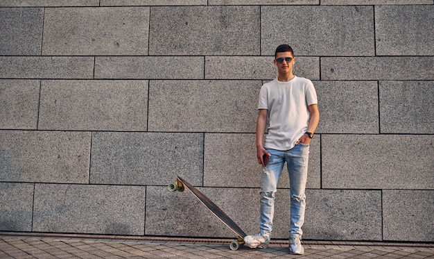 Chico americano caucásico joven en vasos con un longboard contra la pared gris de un edificio urbano