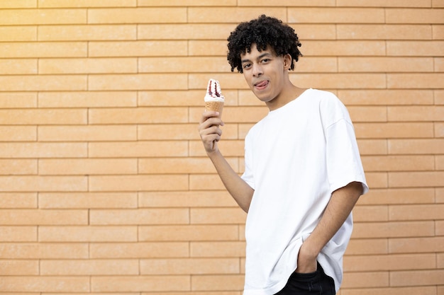 Chico americano rizado sonriente sosteniendo un cono de helado en la mano y lamiendo sus labios