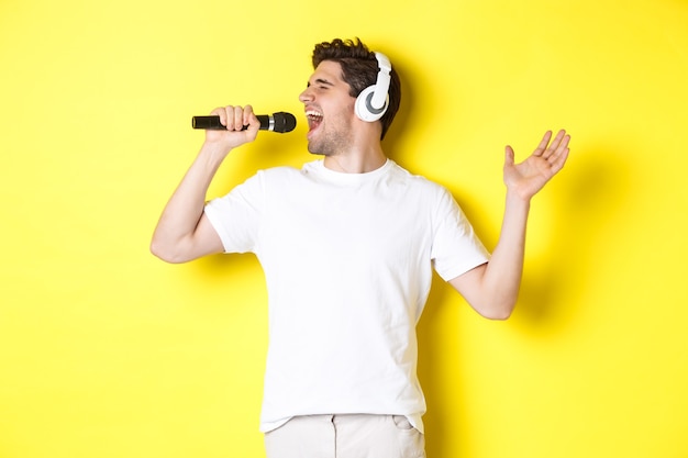 Chico apasionado en auriculares con micrófono, cantando canciones de karaoke, de pie sobre una pared amarilla con ropa blanca.