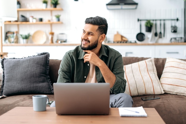 Chico árabe atractivo y positivo con ropa informal sentado en un sofá en casa en la sala de estar usando una computadora portátil navegando por Internet aprendiendo seminarios web de escucha remota mirando hacia otro lado sonríe soñando