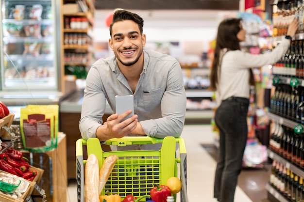 Chico árabe con compras de teléfonos inteligentes de pie con carrito en el supermercado