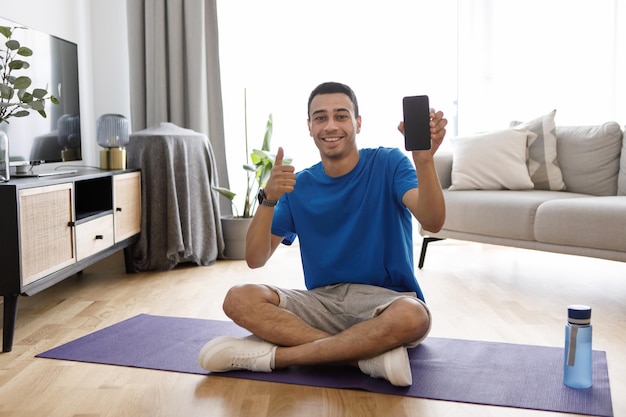 Chico árabe positivo sentado en una alfombra de yoga demostrando un teléfono inteligente con pantalla en blanco y mostrando una maqueta con el pulgar hacia arriba