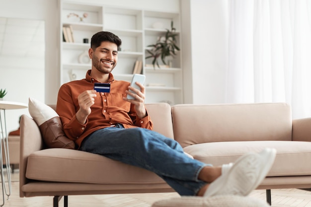 Chico árabe sonriente usando teléfono y tarjeta de crédito en casa