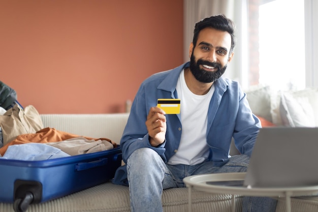 Chico árabe con tarjeta de crédito sentado cerca de una laptop en casa