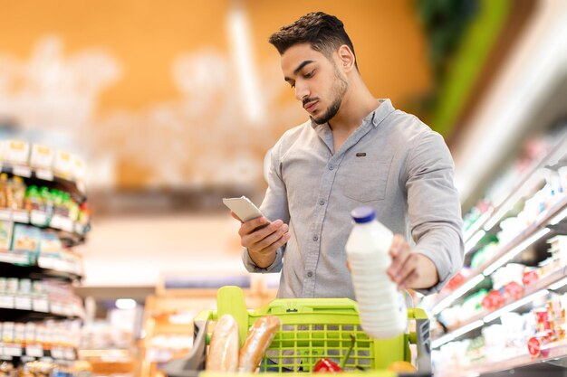 Chico árabe usando teléfono celular sosteniendo leche comprando comestibles en el supermercado