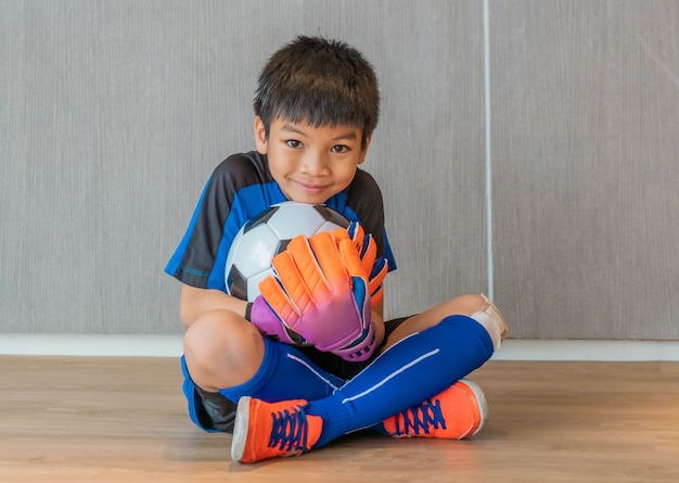 Chico asiático es un portero de fútbol con guantes y sosteniendo un balón de fútbol con cara de sonrisa.