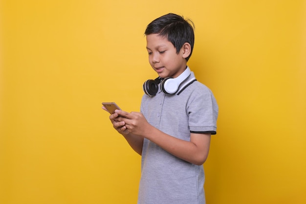 Un chico asiático con estilo informal sonríe mientras usa un teléfono inteligente y auriculares aislados en un fondo amarillo