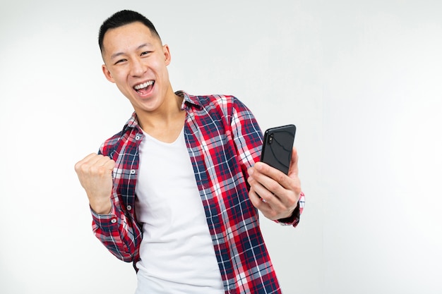 Chico asiático feliz ganó un premio mientras sostiene un teléfono inteligente sobre un fondo blanco.