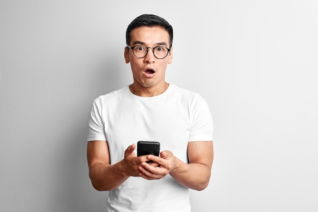 Un chico asiático guapo en un estudio blanco se ve sorprendido por la pantalla del teléfono móvil. Sorprendido hombre kazajo con anteojos vestido de manera informal sostiene el teléfono inteligente en las manos