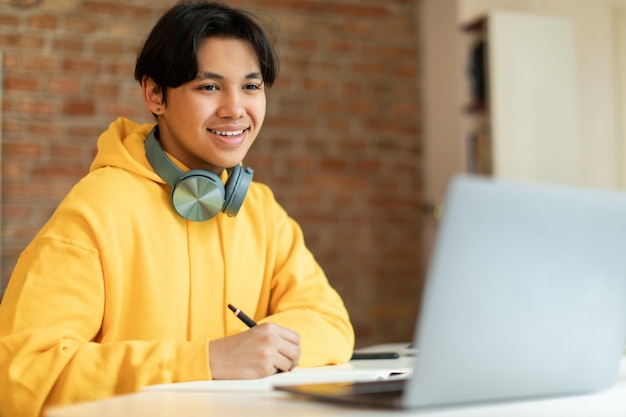 Chico asiático mirando la computadora portátil haciendo la tarea escolar en línea en el interior