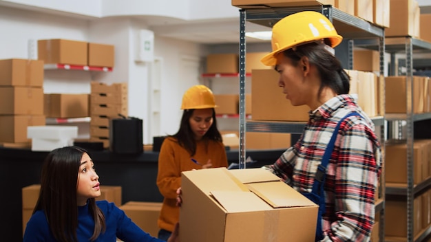 Chico asiático organizando cajas con suministros en estantes, haciendo control de calidad con empresaria en almacén. Empleado poniendo paquetes en estantes, preparándose para la gestión de pedidos.