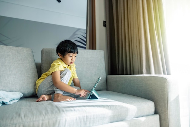 Un chico asiático con el pelo negro y una camisa amarilla está sentado en una tableta Disfrutando en el sofá gris de la sala de estar junto a la ventana