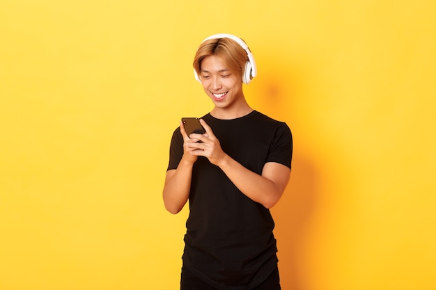 Chico asiático sonriente joven que usa el teléfono inteligente y escucha música o podcast en auriculares inalámbricos