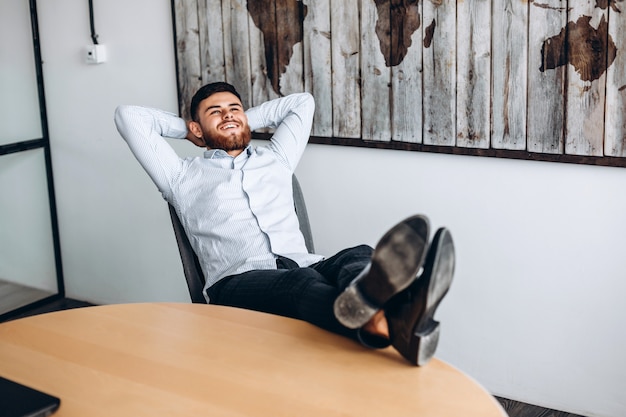 Un chico atractivo con barba pone las manos detrás de la cabeza, pone los pies sobre la mesa y descansa.