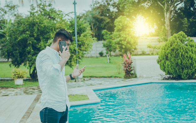 Chico atractivo de pie hablando por teléfono cerca del borde de la piscina Hombre guapo llamando por teléfono junto a una piscina Hombre joven de pie llamando por teléfono junto a una piscina