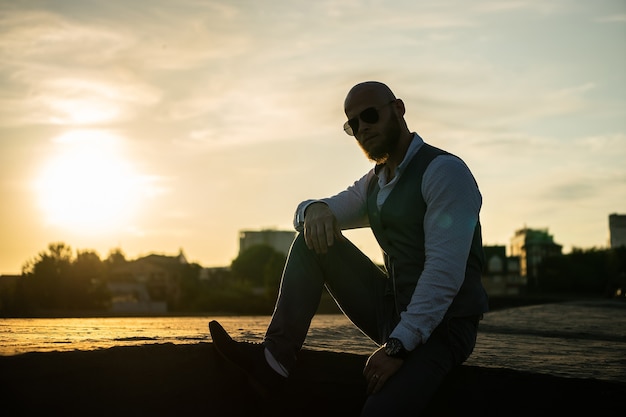 Chico audaz con una elegante barba y gafas de sol en una ciudad borrosa durante la espectacular puesta de sol. Concepto de éxito y voluntad