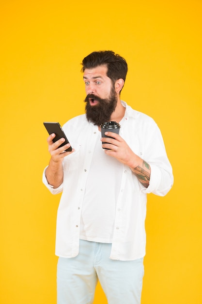 Chico con barba y bigote mantenga teléfono móvil. comunicación. rutina de la mañana. El hombre barbudo sorprendido tiene una llamada telefónica entrante mientras bebe café. brutal hipster bebiendo bebida para llevar de la taza.