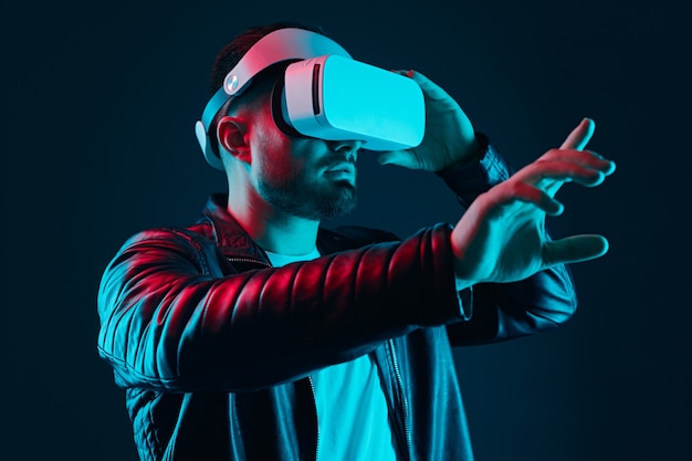 Chico barbudo con chaqueta de cuero negro y gafas de realidad virtual interactuando con la pantalla invisible mientras experimenta la realidad virtual en un estudio oscuro con luces de neón