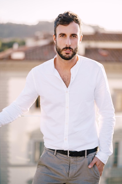 Chico barbudo elegante con una camisa blanca y pantalones claros en una terraza en la azotea en florencia, italia